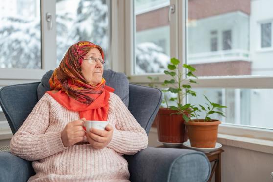 An older woman holding a coffee mug, sitting on a couch, staring out of a window